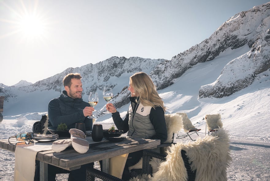 Frau mit blonden Haaren und Mann mit kurzen braunen Haaren sitzen auf Terrasse und trinken Wein